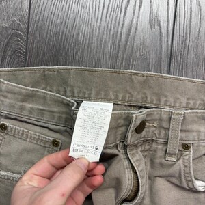 Puede incluir: Primer plano de unos vaqueros Carhartt color beige. Los vaqueros tienen cierre de bot&oacute;n y cremallera, bolsillos delanteros y trabillas para el cintur&oacute;n. Se ve una etiqueta blanca con texto. Los vaqueros son de la talla 32x30.