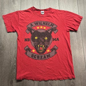 Puede incluir: Una camiseta roja de manga corta con un gráfico de una cabeza de pantera negra con ojos amarillos y dientes al descubierto. El diseño incluye el texto A WILHELM SCREAM, las letras NB y MA, y cinco estrellas amarillas en un diseño circular.