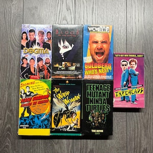 Może przedstawiać: Kolekcja starych kaset VHS z tytułami filmów, takimi jak "Dogma", "Blood: The Last Vampire" i "Teenage Mutant Ninja Turtles: The Movie". Inne tytuły to "Tapeheads" i "The Wasp Woman". Kasety są ułożone na drewnianej powierzchni.
