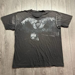 May include: A charcoal gray short-sleeve t-shirt featuring a distressed white graphic of a muscular figure with arms outstretched. The text UNIVERSAL STUDIOS is printed in white on the lower right side of the graphic. The shirt has a crew neckline.