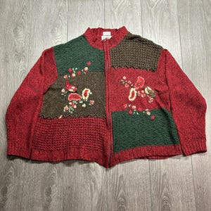 Pode incluir: Um casaco de malha vermelho, verde e castanho com fecho de correr, gola alta e design de patchwork. A peça de malha apresenta motivos florais e de pássaros bordados nos painéis frontais, com secções texturizadas e fecho frontal completo.