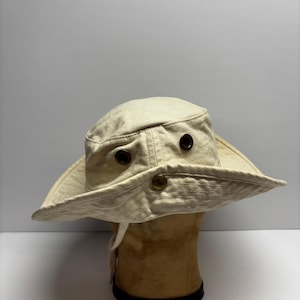 May include: A cream-colored bucket hat featuring a wide, downward-sloping brim, two dark brown metal ventilation eyelets on the side, and a single brass-toned snap button on the front brim. The hat is constructed from a sturdy, woven fabric.