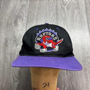 Può includere: Un cappellino da baseball nero con visiera viola caratterizzato dal logo ricamato dei Toronto Raptors sul davanti. Il logo raffigura un dinosauro rosso che tiene un pallone da basket con la scritta Toronto Raptors arcuata sopra in bianco e viola.