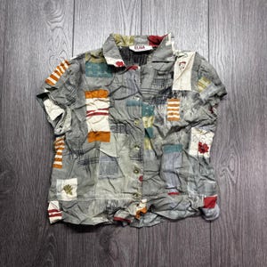 Puede incluir: Una camisa de manga corta con un diseño de patchwork. La camisa presenta una variedad de colores, incluyendo gris, naranja, blanco y rojo. El cuello tiene el nombre de la marca "ELISA". La camisa está hecha de tela arrugada.