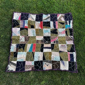 May include: A square patchwork quilt featuring a grid of multi-coloured fabric squares in shades of olive green, black, white, and blue. The quilt has a visible quilted texture throughout and is finished with a solid dark purple fabric border.