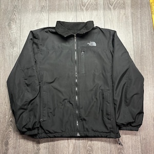 May include: Black jacket with a full-length zip, a small chest pocket, and a fleece-lined collar. The North Face logo is visible on the upper left chest. The jacket has two side pockets and drawstrings at the bottom hem.