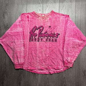 May include: A long-sleeve crewneck shirt in a vibrant pink acid-wash pattern. The front features a black graphic with the text Pro Performance BODY GEAR and a small illustration of a muscular figure flexing an arm above the lettering.