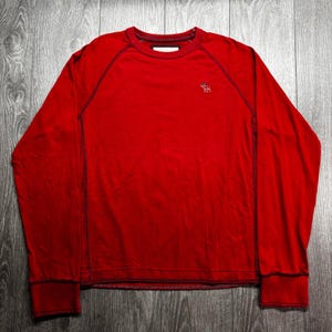 May include: A long-sleeved red crew-neck shirt featuring dark contrast stitching along the raglan sleeves and hem. A small white embroidered moose logo is positioned on the upper left chest area. The garment has a ribbed collar and cuffs.