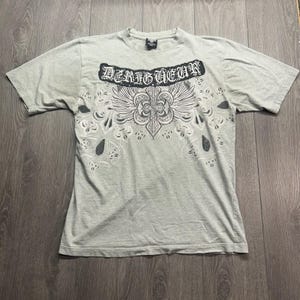 May include: A light gray short-sleeve t-shirt featuring a black graphic print on the chest. The design includes the text "DERIGUEUR" in a gothic font above a stylized cross with wings and intricate scrollwork patterns extending downwards.