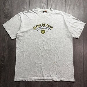 Può includere: Una t-shirt a maniche corte grigio chiaro con una grafica centrale che riporta il testo ESPRIT DE CORP. FASHION NOUVEAU SAN FRANCISCO, CA Est. 1968 attorno a un emblema a stella verde e giallo. La maglia presenta un girocollo e tessuto mélange.