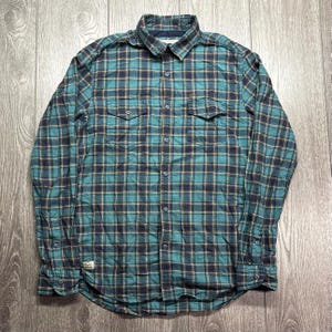 May include: A teal, navy, and tan plaid button-down shirt. The long-sleeved shirt features two chest pockets and a button-down collar. The fabric has a quilted appearance. The shirt is displayed on a wooden surface.