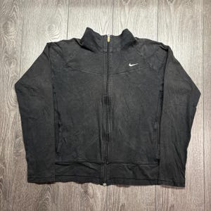 May include: A black zip-up jacket with a high collar and a small white Nike logo on the chest. The jacket has a full-length zip and two side pockets. The fabric appears to be a faded, textured material.
