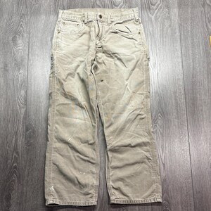 Puede incluir: Pantalones de trabajo color caqui con m&uacute;ltiples bolsillos y cierre de bot&oacute;n y cremallera. Los pantalones muestran signos de desgaste, con algunas marcas de pintura y manchas. Los pantalones est&aacute;n hechos de un material duradero.