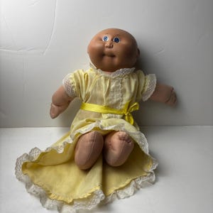 Puede incluir: Muñeca Cabbage Patch Kid vintage con cabeza calva y ojos azules. La muñeca lleva un vestido amarillo con ribete de encaje blanco y una cinta amarilla. La piel de la muñeca es de color marrón claro.