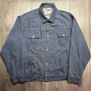 May include: A light blue denim jacket with a classic collar and button-down front. The jacket has two chest pockets and two side pockets. It features silver buttons and visible stitching. The jacket is laid flat on a wooden surface. The jacket is a vintage item.