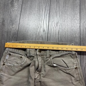 Puede incluir: Primer plano de un pantal&oacute;n de trabajo gris descolorido con una regla de madera colocada sobre la cintura. El pantal&oacute;n tiene cierre de bot&oacute;n, bolsillos delanteros y trabillas. La regla mide hasta 43 cm.