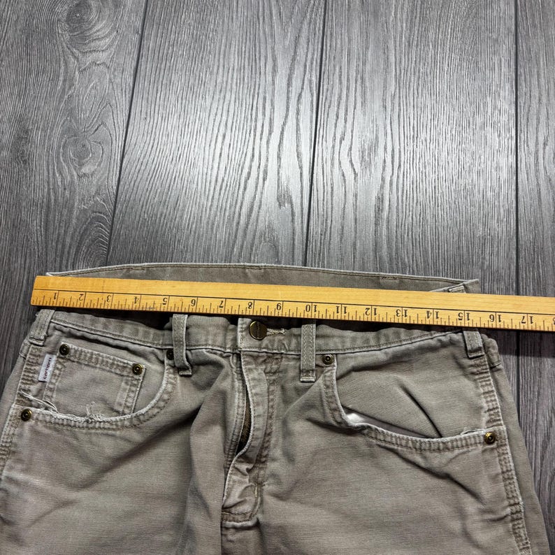 Puede incluir: Primer plano de un pantal&oacute;n de trabajo gris descolorido con una regla de madera colocada sobre la cintura. El pantal&oacute;n tiene cierre de bot&oacute;n, bolsillos delanteros y trabillas. La regla mide hasta 43 cm.