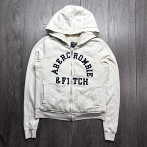 May include: A cream-coloured zip-up hoodie featuring a sherpa-lined hood and dark navy blue lettering across the chest that reads Abercrombie & Fitch. The garment includes long sleeves, a front zipper closure, and two front pockets.