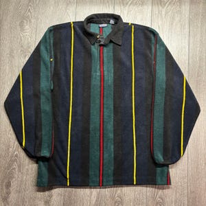 Peut inclure: Un pull en polaire à manches longues avec un motif à rayures verticales bleu marine foncé, vert forêt et noir. Le vêtement présente un col, une patte de boutonnage partielle et de fines rayures verticales accentuées en jaune et rouge.