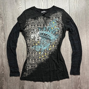 May include: A black long-sleeve shirt featuring a diagonal panel with a grey damask pattern. The design includes a light blue crown graphic, a white fleur-de-lis, and small yellow stars scattered across the front of the garment.