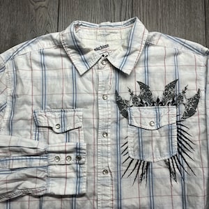 May include: White, blue, and red plaid button-up shirt with long sleeves. The shirt has two front pockets and a black graphic design on the front. The word "machine" is printed on the collar.
