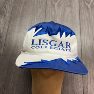 Puede incluir: Una gorra de béisbol blanca y azul con un patrón bordado en zigzag tipo rayo a lo largo de la corona y la visera. El panel frontal muestra el texto bordado en azul LISGAR COLLEGIATE en una fuente serif centrada sobre la tela blanca.