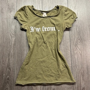 May include: An olive green short-sleeved t-shirt featuring a wide scoop neckline with a small white ribbon detail at the centre. The front of the shirt displays the white text "I'm from." in a distressed, gothic-style font across the chest.