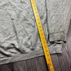 Puede incluir: Una sudadera gris con cuello redondo y pu&ntilde;os acanalados. Una regla de madera amarilla se coloca en diagonal sobre la sudadera, mostrando medidas en cent&iacute;metros. La sudadera est&aacute; sobre una superficie de madera oscura.