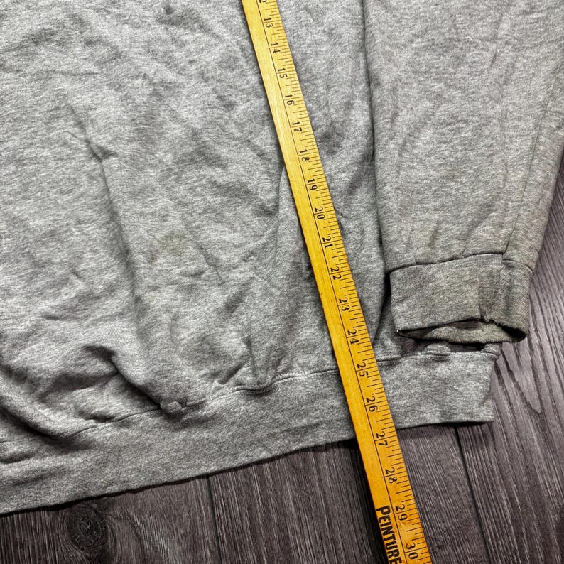 Puede incluir: Una sudadera gris con cuello redondo y pu&ntilde;os acanalados. Una regla de madera amarilla se coloca en diagonal sobre la sudadera, mostrando medidas en cent&iacute;metros. La sudadera est&aacute; sobre una superficie de madera oscura.