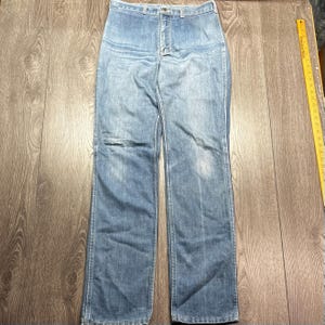 May include: Light blue denim jeans with a button closure and a classic five-pocket design. The jeans have a straight leg cut and show signs of wear. The garment is laid flat on a wooden surface.