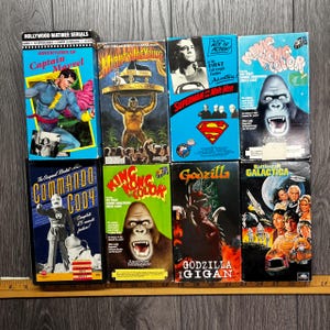 May include: A selection of retro VHS tapes showcasing classic films and serials. Titles include "Captain Marvel," "Mighty Joe Young," "Superman and the Mole Men," "King Kong," "Commando Cody," "Godzilla vs. Gigan," and "Battlestar Galactica." The tapes feature vibrant cover designs.
