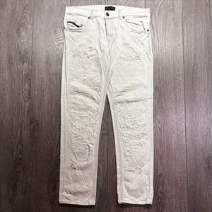 May include: Off-white denim jeans featuring a heavily distressed and ripped texture across the front panels. These trousers include a standard button and zip fly closure, two front pockets, a small coin pocket, and belt loops along the waistband.