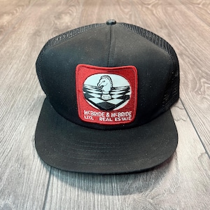 Puede incluir: Gorra de camionero negra de malla con un parche bordado en rojo y blanco que presenta una pieza de ajedrez y el texto "McBride & McBride LTD. Real Estate".