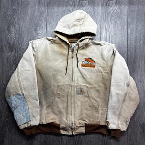 Peut inclure: Une veste à capuche zippée beige avec un ourlet et des poignets côtelés marron. La poitrine présente un écusson brodé avec un graphique de camion orange et le texte Mason County Road Commission. La veste montre des signes d'usure et des rapiéçages.