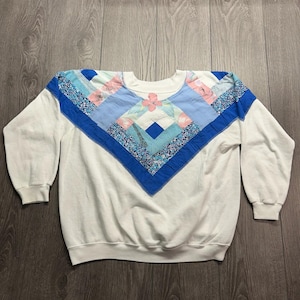 May include: A white long-sleeve sweatshirt featuring a V-shaped patchwork design across the chest and shoulders. The patchwork consists of various blue, light blue, and pink floral fabric squares arranged in a geometric pattern with a crew neck.