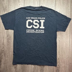 May include: Dark blue t-shirt with the text "LAS VEGAS POLICE CSI CRIME SCENE INVESTIGATOR" printed in white. The shirt is a crew neck and appears to be made of cotton. The shirt is slightly faded.