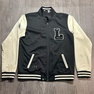 Peut inclure: Une veste zippée noire avec des manches couleur crème, dotée d'un col montant et de poignets côtelés à rayures noires et blanches. La poitrine présente un grand écusson en forme de lettre L avec le texte LRG CLASS OF 47 et le nombre 1947 brodé.