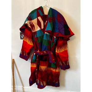 Puede incluir: Una bata colorida con estampado y cinturón, colgada de una percha metálica. La bata presenta un diseño geométrico en rojo, azul, verde y amarillo. La bata está hecha de un material grueso y el cinturón está atado a la cintura.