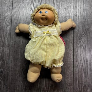 May include: A vintage Cabbage Patch Kid doll with blonde yarn hair and blue eyes. The doll is wearing a yellow romper with a white and yellow checkered pattern, lace trim, and a yellow bow. The doll's arms and legs are outstretched.