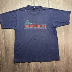 May include: A navy blue short-sleeve t-shirt featuring a graphic of a loon swimming in water with green reeds. The text NORTHERN REFLECTIONS is printed in red block letters with a white outline centred on the chest of the garment.