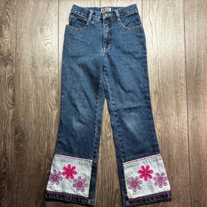May include: High-waisted blue denim jeans featuring a straight-leg cut with light-wash denim cuffs at the bottom. The cuffs are decorated with embroidered pink and purple flower motifs. The jeans include a metal button closure and a Request brand label.