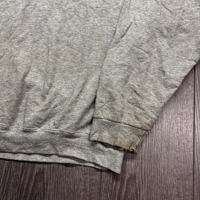 Puede incluir: Primer plano de una manga de sudadera gris. El pu&ntilde;o est&aacute; desgastado y descolorido. La tela es gris jaspeado, mostrando signos de desgaste y uso. La sudadera parece estar hecha de un material suave y c&oacute;modo.