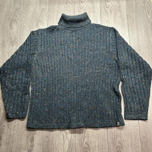 Op de afbeelding: Een donkerblauwe coltrui met een verticaal geribbeld breipatroon en een veelkleurig gespikkeld ontwerp. Het kledingstuk heeft lange mouwen en een klein rechthoekig logolabel bij de onderste zoom aan de voorzijde.