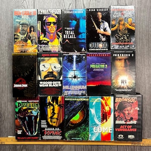 May include: A selection of vintage VHS tapes, including titles such as "Iron Eagle III," "Total Recall," and "Jurassic Park." The tapes display vibrant movie artwork and titles. The collection is arranged on a wooden surface, showcasing classic film covers.