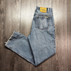 Puede incluir: Vaqueros de mezclilla azul claro con un diseño clásico de cinco bolsillos. Los vaqueros presentan un lavado descolorido y un parche de cuero con el nombre de la marca. Los vaqueros están colocados sobre una superficie de madera.