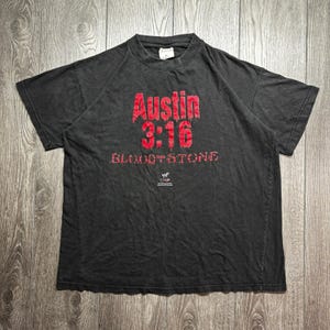 Peut inclure: Un t-shirt noir à manches courtes avec un texte rouge indiquant Austin 3:16 et BLOOD FROM STONE. Sous le texte se trouve un petit logo WWF blanc. Le t-shirt a un col rond et une coupe standard, avec un motif graphique imprimé sur la poitrine.