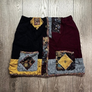 Op de afbeelding: Een patchwork minirok in zwart, bordeauxrood en grijs. Het heeft decoratieve zakken met gouden en patroonaccenten en knopen aan de voorkant. De rok is gemaakt van een zachte, getextureerde stof. Het ontwerp is een mix van texturen en patronen.