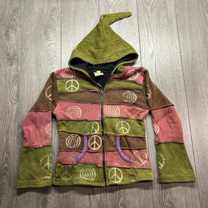 Könnte beinhalten: Ein langärmeliger Kapuzenpullover mit Reißverschluss und Patchwork-Design in Olivgrün, Braun und gedämpftem Rot. Das Kleidungsstück verfügt über eine spitze Kapuze, zwei Vordertaschen und weiße Siebdruckmotive von Friedenszeichen und Spiralen.