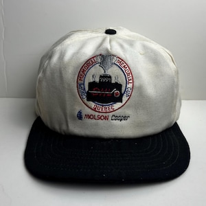 May include: A white and black baseball cap with embroidered text and graphics. The cap features the words "Coupe Memorial Cup" in a circular design, a trophy graphic, and the text "Quebec" and "Molson Cooper".