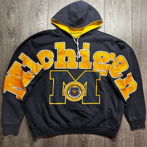 Peut inclure: Un sweat-shirt à capuche bleu marine avec le mot Michigan en grandes lettres jaunes sur la poitrine et les manches. Une grande lettre M jaune est centrée avec le sceau de l'Université du Michigan et le texte 1817 imprimé dans l'emblème.